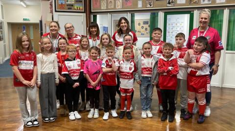 Staff and pupils at Spring Cottage Primary School stand in a school hall. The floor is shiny, dark wood. The children are wearing red and white rugby league shirts and are surrounded by adults wearing the same clothing. Drawings by the pupils are also on the walls.