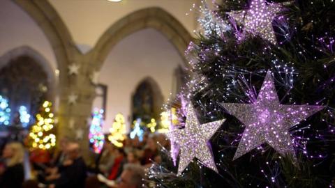 The side of a Christmas tree with silver stars on it and a blurred background of people at a carol service in a Jersey church.