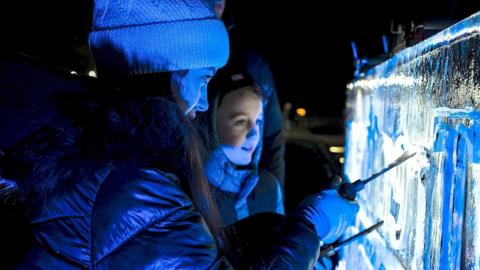A girl in a black hooded coat and woolly hat marking something into an ice sculpture in front of her with another child stood next to her smiling