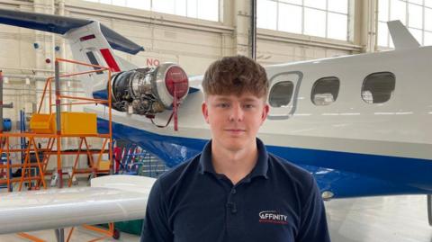 A young man with brown hair draped over his forehead who is clean shaven is smiling at the camera and wearing a navy polo shirt which says Affinity on it. Behind him is a small aircraft which is white, blue and red, which is stationary inside a large warehouse building.