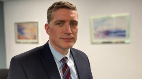 A man with short, light coloured hair, staring at the camera. He is wearing a blue suit jacket, a light blue shirt and a red and blue striped tie.  A blurred white wall with artwork is in the background.
