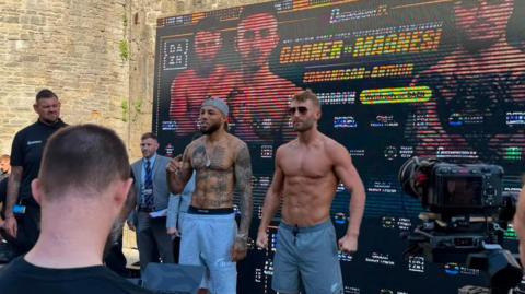 Ryan Garner (right) Michael Magnesi (left). Both men are stood on a stage in shorts with no tops on. They standing in fighting poses and looking out to the crowds, while there is a board behind them advertising the fight. Garner is wearing sunglasses.