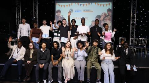 A group of young people sit and stand on a stage as they smile and wave at the camera celebrating. Behind them is a screen and a number of silver chairs.