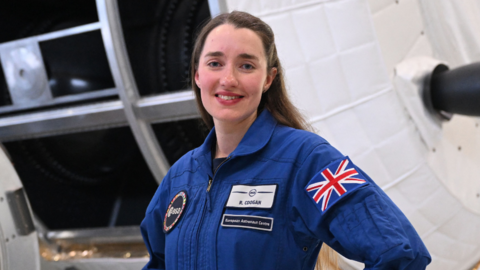 Rosemary Coogan wearing a European Space Agency blue jump suit, has her hands on her hips and is smiling towards the camera. She is stranding in front of two large spacecraft.