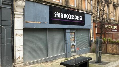 A shop front on an urban street. The shop name 'Sasa Accessories' is above the windows. Shutters are down within the shop. A small marble bench sits outside.