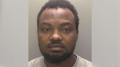 A police custody picture of a man with black hair and a cropped black beard and moustache. He is wearing a light-grey T-shirt and has an emotionless expression.