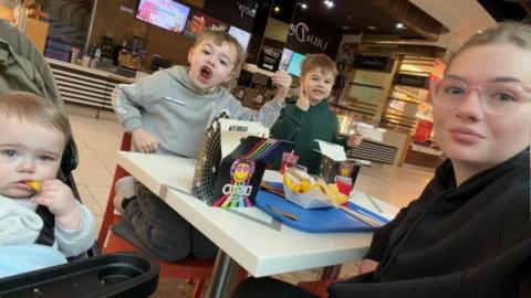 A woman is taking a selfie of her family in a fastfood restaurant - she is on the right of the table and has a black top on with her hair tied back and pink rimmed glasses on. Her two sons are in the middle, eating from a little black cardboard box of food and looking straight at the camera. Her daughter is to the left, she is a baby of around a year old and is eating a chip in a pushchair, looking at the camera.