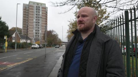 Michael Jones, who is bald and has a short ginger beard, is standing outside Willow Rise in Kirkby. The building is a 1960s tower block. Mr Jones is looking at a reporter who is off camera. 