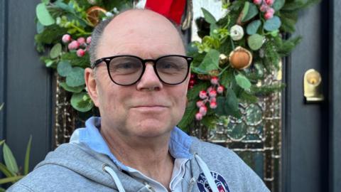 Resident John Lyons, wearing a grey Bath Rugby hoodie, stands smiling by his front door, which has a wreath on it.