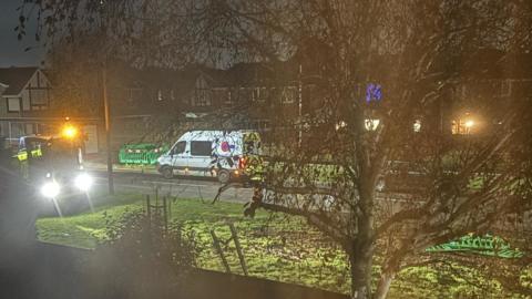 A view over a garden wall of vans parked up on a roadside. Their lights are on and it's dark.