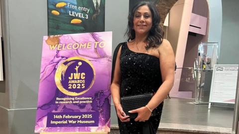 A woman with dark hair is wearing a one-shoulder sparkly black dress. She is standing indoors in front of some marble steps. She is standing next to a purple sign that says "welcome to JWC awards 2025"