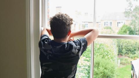 A teenage boy looking out of a window and onto a house's back garden, with terraced houses in the background 