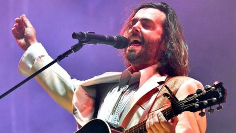 Lord Huron singer Ben Schneider on stage in a white suit with mouth open and eyes closed mid song, with one hand on a guitar neck and the other in the air. At the 2022 Bonnaroo Music & Arts festival in Tennessee.