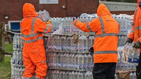 Two people in orange high vis jackets. They are facing a large crate of bottled water.