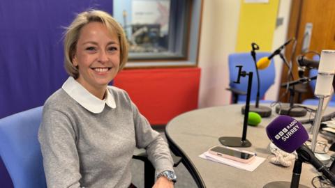 Lisa Townsend in a grey top, smiling at the camera. She is sat in a radio studio with a purple microphone branded BBC Radio Surrey in front of her.