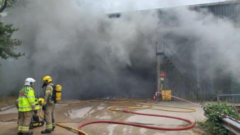 Two firefighters stand outside a building that is clouded in smoke. 