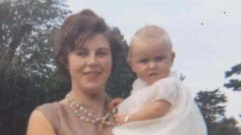 An old photo of Sandie Bowen as a young mum, holding her baby daughter Anita in her arms. Sandie is wearing a pink dress and colourful bead necklace and has short brown hair. Baby Anita is in a white Christening dress.