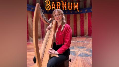 A young woman with dark blonde hair is sitting on a stool on a theatre stage with an American flag type background. She s wearing a red denim cropped jacket, white t-shirt and black jeans and is holding a large light brown harp with colourful strings. On the stage backdrop is the word "Barnum" in large letters lit up by bulbs