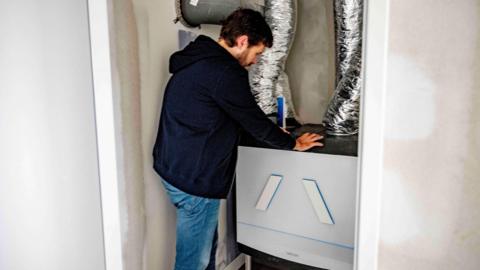 A man is resting his hands on a boiler which has insulated tubes leading upwards. He is wearing jeans and a navy hoodie.