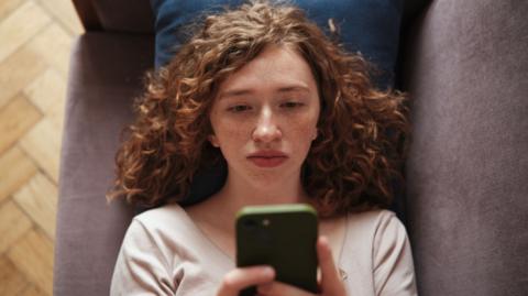 A woman lying on a sofa holding her phone up looking bored