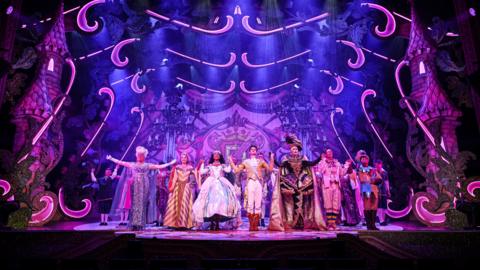 A photo of a stage with a pantomime cast about to take their bows. The set is purple and has castle turrets on either side of the cast.