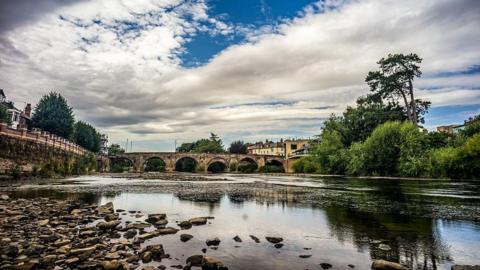 The river bed of the River Wye is seen exposed looking toward Old Bridge after weeks of no rainfall. Buildings and trees can be seen along the right-hand-side of the bank.