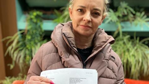 A blonde woman wearing a taupe-coloured coat is holding a document.