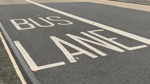 Image shows the words 'BUS LANE' painted in large white capital letters on the road surface by a kerbside.
