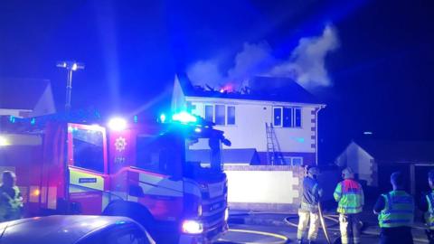 Flames and smoke rise from a collapsed roof on a house. It is night time and the scene is illuminated by the flashing blue lights of a fire engine parked in front of the two storey property.