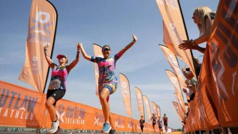 Two athletes at the finish line of the Nottingham Outlaw Triathlon. They are running on an orange carpet with spectators looking over an orange barrier cheering on the two female runners. The two women have their arms in the air and are holding hands.