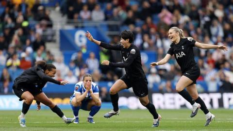 Three women footballers in black kit celebrate on the pitch while a player in blue kit sits on her haunches with her hands to her mouth. 