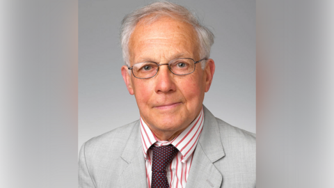 A portrait of a man with short white hair, glasses, a dark red spotted tie, a red and white shirt and a grey jacket.
