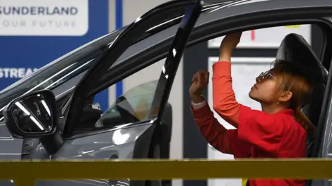 A woman in a red jumpsuit inside a car in a factory, reaching arm up to the roof and inspecting it.