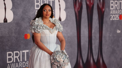 British singer Jade poses on the red carpet during the 2025 BRIT Awards ceremony at The O2 arena in London. She wears a denim structured dress with ruched flowers on her shoulders and is holding a denim bouquet with both hands.