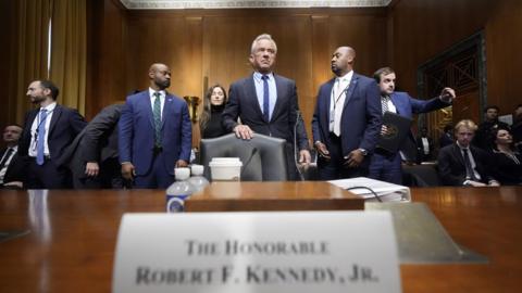 Robert F Kennedy Jr stands behind a chair, pushed up to a wide, wooden table. He is wearing a smart, tailored, dark suit. He is flanked by other similarly dressed men. On the table is a sign with his name on it, some plastic bottles and a white disposable drinks cup. 
