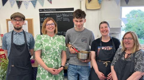 Five people stand in front of a blackboard. Jenny Lane who has re-started Workbridge wears a green and white patterned dress. A young man next to her holds a saucepan containing spagetti bolognaise.