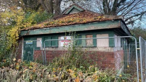 The old toilet block is a one story brown-brick building with a triangle roof and green door and window panes. There is a pile of yellow tree leaves on the roof. NO UNAUTHORISED ACCESS is written in red on a sign attached to the building. A grey metal fence is surrounding the block.