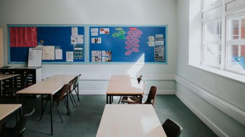 A stock image of a classroom, with desks and chairs behind them, with a wall displays at the back of the room and windows to the right of the picture.