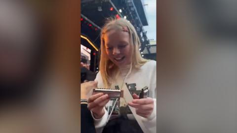 A blonde girl in a white jumper holds a harmonica at a concert