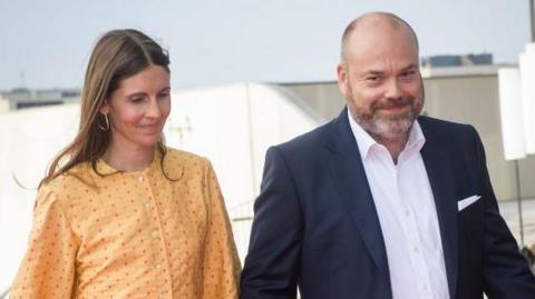 The husband and wife are smiling while they walk. There are buildings in the background.