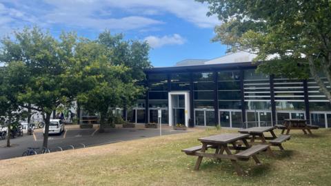 A black building, walled with dark glass. In the foreground is grass with 3 picnic tables on it. There are trees at either end of the picture.