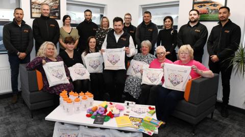 A group of employees from Inco are standing up in black uniforms with an orange Inco logo. In front of them on a sofa are six women from the hospital trust who are holding the care bags. They have a heart on the front. In the middle of the women is Tim Lewis, who is also holding a care bag.