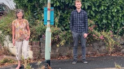 Roberta Ayles on the left in a flowery top and Ethan Merrington-Pink on the right in a check shirt with a bus-stop between them 