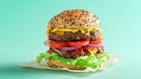 A beef burger in a seasame seed topped bun. The burger layers from bottom to top are bottom of burger bun, lettuce, beef patty, relish, red onion, tomato, beef patty, cheese, burger, top of burger bun. 
