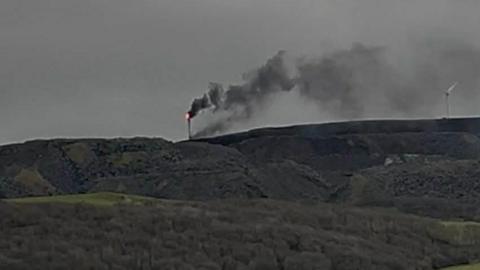 A slightly blurry photo of the wind turbine on fire in the distance on top of a grassy hill. Flames can be seen on the top of the white structure with black smoke coming off it. There is another wind turbine to the right which is not on fire.