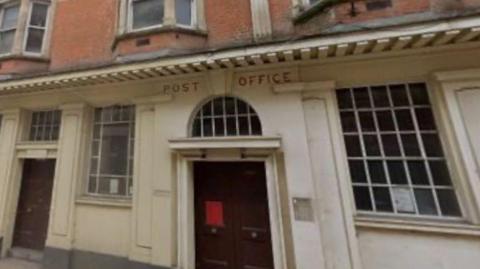 The front of an old post office. The building is white with a brown door. The building says POST OFFICE on it in red letters. There are windows on either side of the main door. To the left of the window on the left of the door is another smaller door. Above the white part of the building is red brick and windows. 