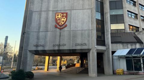 A grey, brutalist concrete building front with "Woking Borough Council" written in letters. There is a gold and red crest on the front of the building.