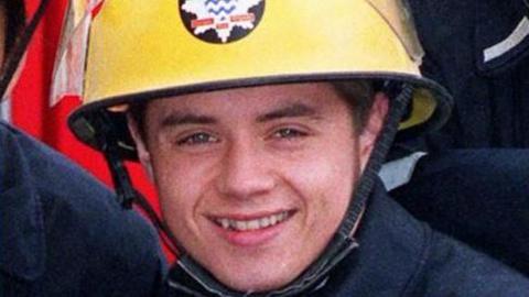 John Alford playing the part of a firefighter. He is wearing a yellow helmet with a star-shaped badge and is smiling.