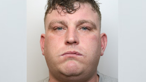 A mugshot of a man in a grey T-shirt looking seriously at the camera. He has brown hair, shaved at the sides and longer on top. It looks messy and wet. He has blue eyes and he looks tired. There is something on his lip, possibly a cold sore.