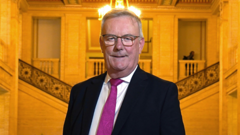 Mike Nesbitt in navy suit and pink tie stood in Stormont great hall